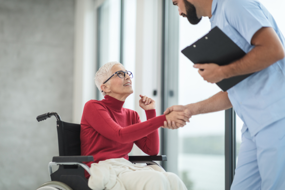 A support worker with a clipboard greeting an older woman in a wheelchair