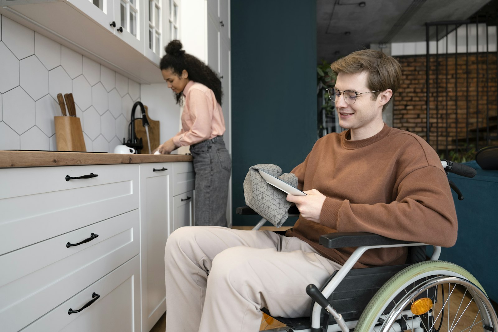 A young man in a wheelchair relaxing in his kitchen while a support worker prepares food