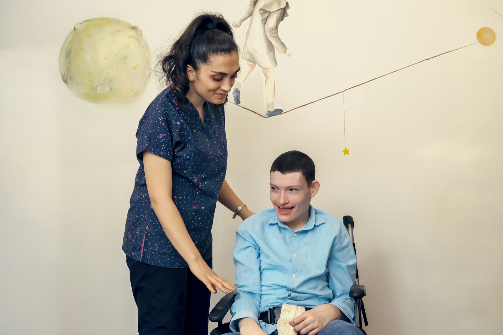 A smiling support worker in scrubs with a young man in a wheelchair, the two of them sharing a laugh