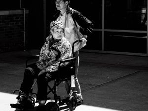 An older woman in a wheelchair, helped by a companion, waiting outside for her transport