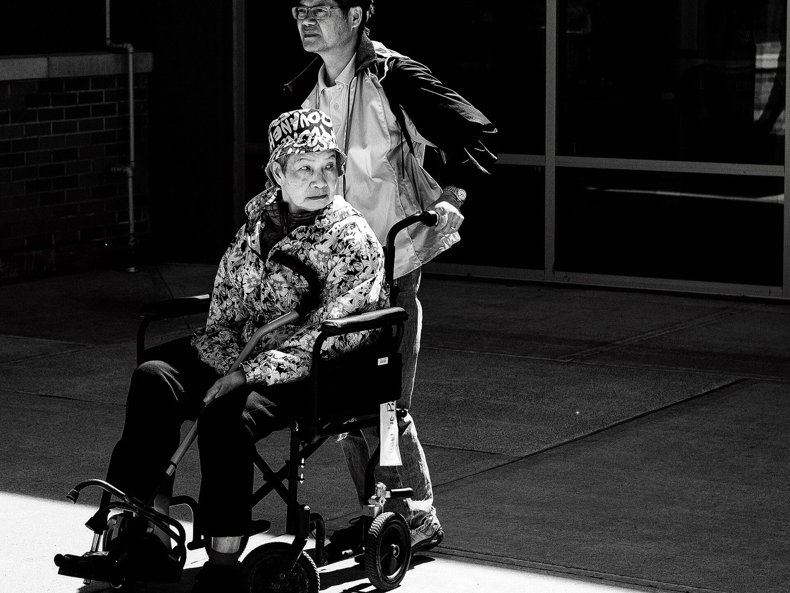 An older woman in a wheelchair, helped by a companion, waiting outside for her transport