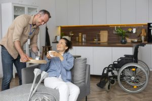 A support worker brings a hot drink to a young woman relaxing on her couch at home, with her wheelchair nearby