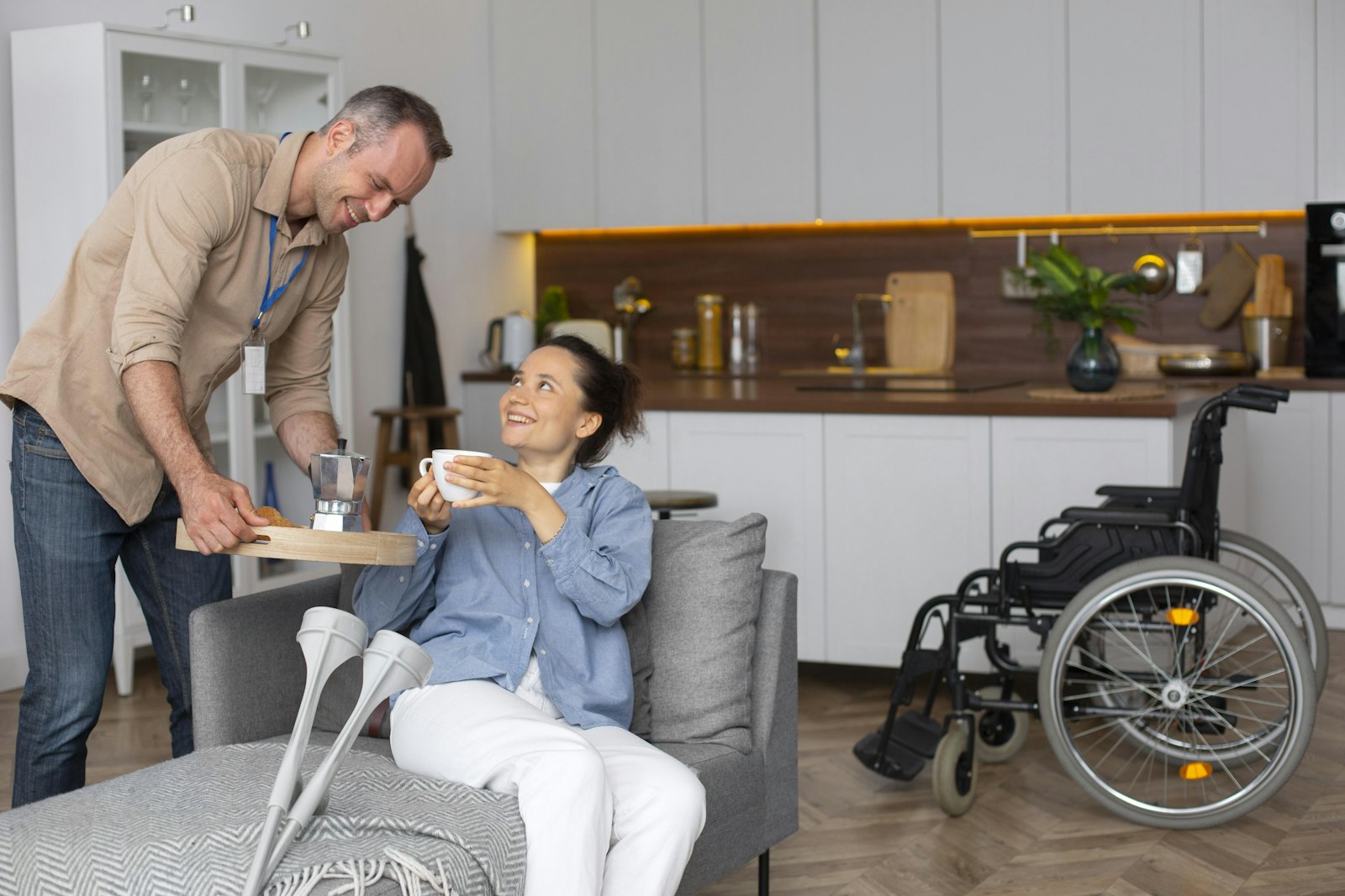 A support worker brings a hot drink to a young woman relaxing on her couch at home, with her wheelchair nearby