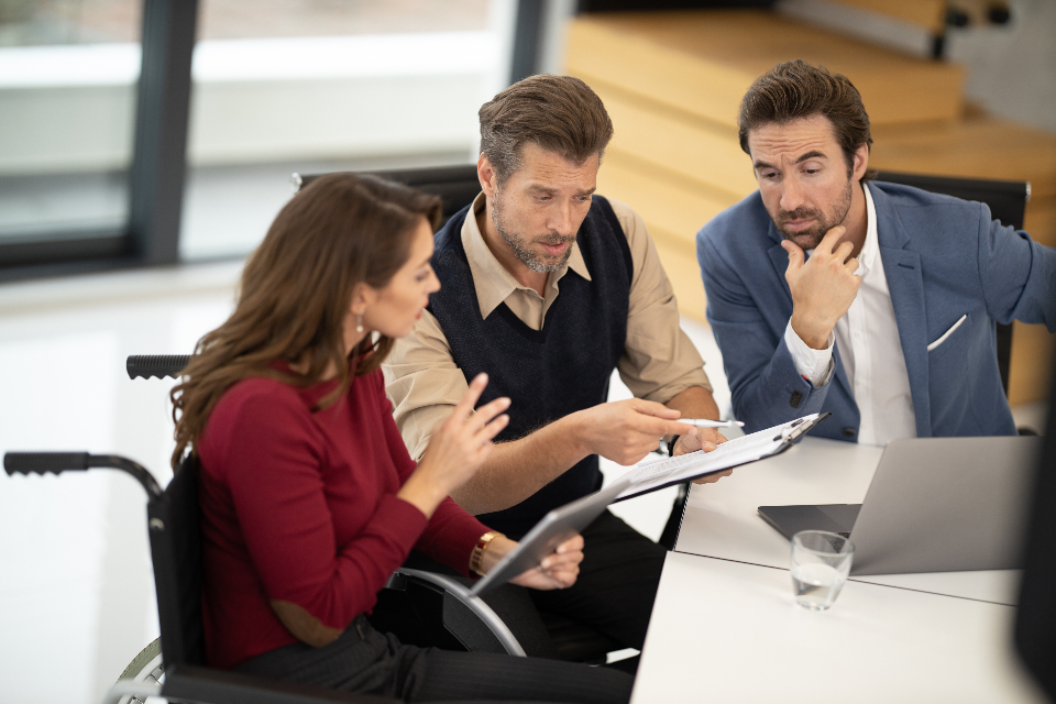 A woman using a wheelchair reviews a printed plan with two colleagues at a table