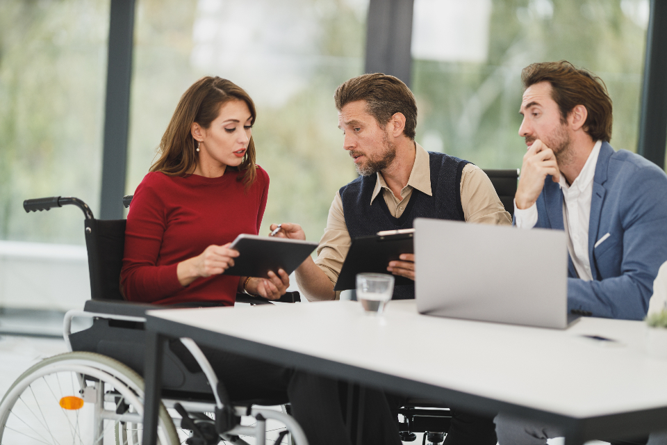 A woman using a wheelchair reviews a plan on a tablet with two colleagues at a meeting table